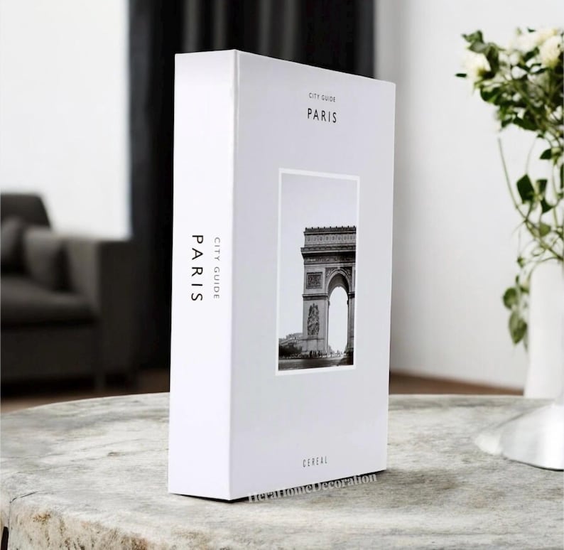 K&ouml;nnte beinhalten: Eine wei&szlig;e rechteckige Box mit den Worten "CITY GUIDE PARIS" auf der Vorder- und Seite aufgedruckt. Auf der Vorderseite befindet sich ein Schwarz-Wei&szlig;-Foto des Arc de Triomphe. Das Wort "CEREAL" ist unten aufgedruckt.