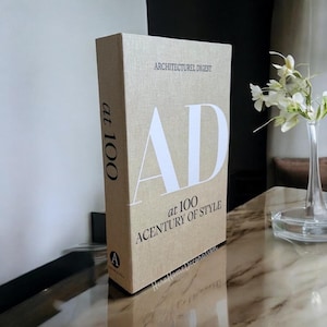 May include: A beige decorative book with the text "ARCHITECTURAL DIGEST" and "AD at 100 A CENTURY OF STYLE" in white. The book is standing upright on a reflective surface.