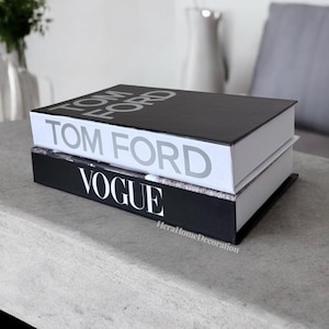 May include: Two hardcover books stacked on top of each other. The top book is black with the words "TOM FORD" in silver. The bottom book is black with "VOGUE" in white. The books are on a light gray surface.
