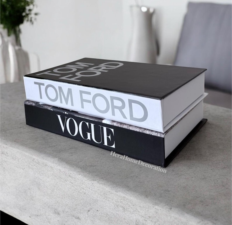 May include: Two hardcover books stacked on top of each other. The top book is black with the words "TOM FORD" in silver. The bottom book is black with "VOGUE" in white. The books are on a light gray surface.