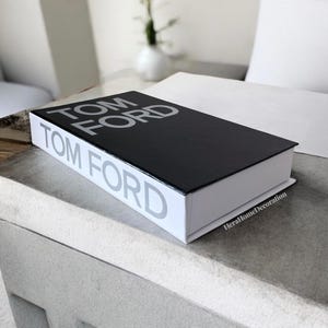 May include: A decorative black and white book with the text "TOM FORD" printed on the cover and spine. The book is rectangular and sits on a light gray surface. The book is a home decor item.