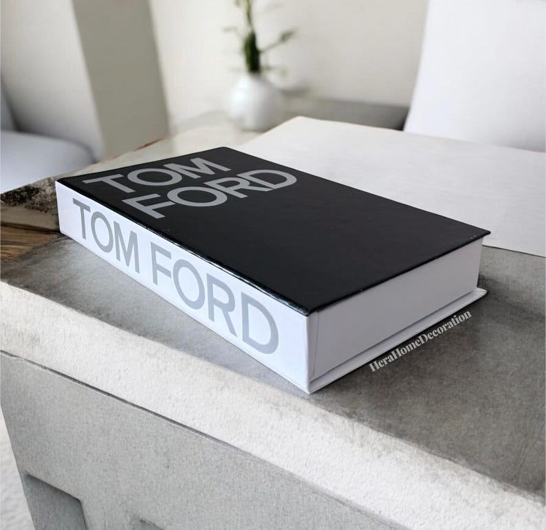 May include: A decorative black and white book with the text "TOM FORD" printed on the cover and spine. The book is rectangular and sits on a light gray surface. The book is a home decor item.