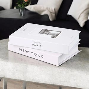 K&ouml;nnte beinhalten: Zwei wei&szlig;e Hardcover-B&uuml;cher, die auf einer hellgrauen Oberfl&auml;che gestapelt sind. Das obere Buch tr&auml;gt die Aufschrift "CITY GUIDE PARIS" mit einem Schwarz-Wei&szlig;-Bild. Das untere Buch tr&auml;gt die Aufschrift "NEW YORK" in fetten schwarzen Buchstaben. Reise- oder Wohndekor-Thema.