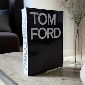 May include: A black decorative book with the text "TOM FORD" in large, silver letters. The spine of the book has the text "TOM FORD" vertically. The book is on a light-colored surface.