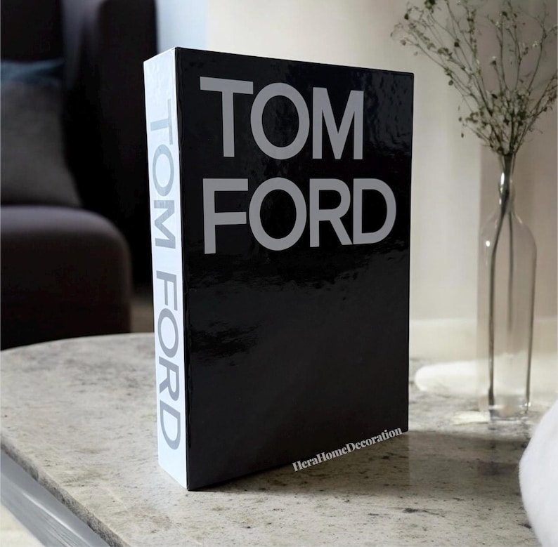 May include: A black decorative book with the text "TOM FORD" in large, silver letters. The spine of the book has the text "TOM FORD" vertically. The book is on a light-colored surface.