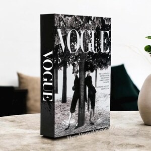 May include: A black decorative book with the word "VOGUE" in white on the spine and cover. The cover features a black and white image of two figures in front of a tree. The book is a home decor item.