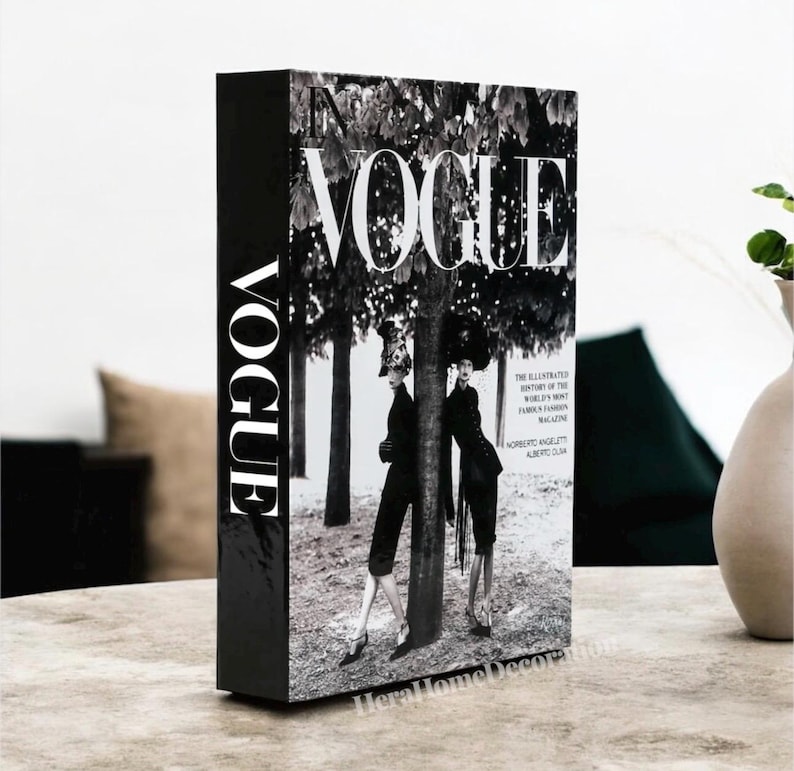 May include: A black decorative book with the word "VOGUE" in white on the spine and cover. The cover features a black and white image of two figures in front of a tree. The book is a home decor item.