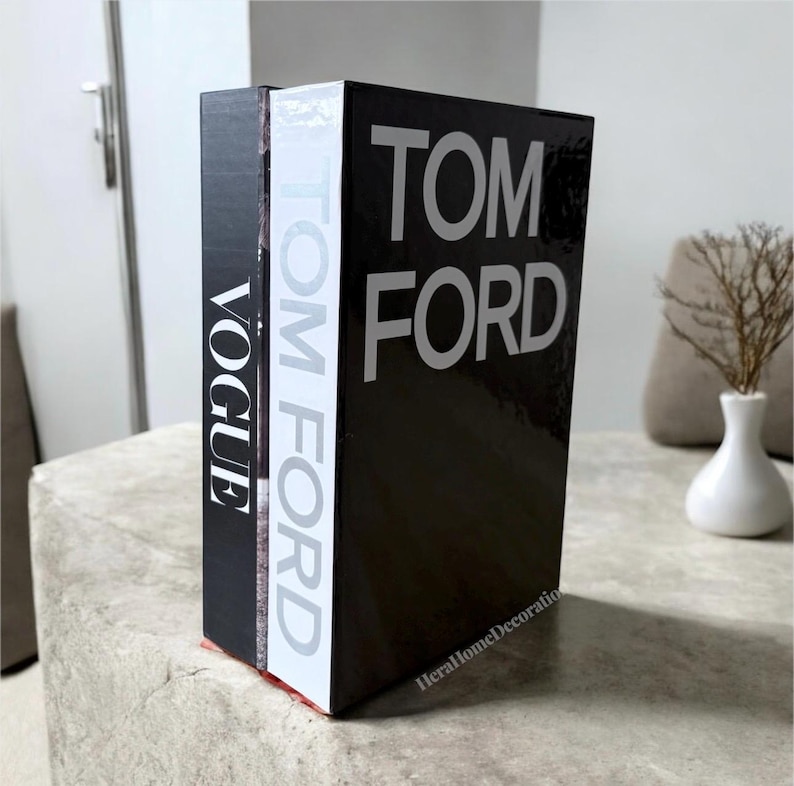 May include: Three decorative books with the titles "VOGUE" and "TOM FORD" in white lettering on a black background. The books are arranged vertically, with the spine of the "TOM FORD" book facing forward. The books are on a light gray surface.