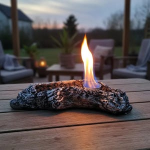 May include: A tabletop fire pit shaped like a piece of textured, weathered driftwood with a dark gray and silver finish. The center contains small dark stones and a circular metal fuel cup with a bright orange and blue flame burning upward.
