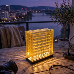 May include: A rectangular glass block lamp with a grid pattern, illuminated with a warm yellow light. The lamp sits on a black base and is plugged into an outlet. The scene is set on a wooden table with a city skyline in the background.