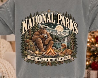 Può includere: Una felpa grigia a girocollo con la grafica di un Bigfoot con occhiali da sole e berretto, seduto accanto a un falò con un cane. Il testo recita NATIONAL PARKS e SAME TRAILS HIGHER SPIRITS, con montagne e un lago.