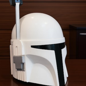 K&ouml;nnte beinhalten: Ein wei&szlig;er Helm mit schwarzen Akzenten, darunter ein T-f&ouml;rmiges Visier und eine vertikale Antenne. Der Helm hat eine graue Seitenkomponente und steht auf einer dunkelbraunen Oberfl&auml;che. Dies ist eine Nachbildung eines Mandalorianer-Helms.