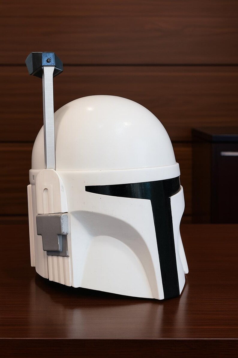 K&ouml;nnte beinhalten: Ein wei&szlig;er Helm mit schwarzen Akzenten, darunter ein T-f&ouml;rmiges Visier und eine vertikale Antenne. Der Helm hat eine graue Seitenkomponente und steht auf einer dunkelbraunen Oberfl&auml;che. Dies ist eine Nachbildung eines Mandalorianer-Helms.