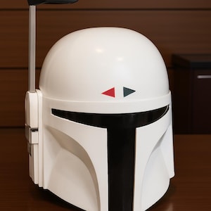 K&ouml;nnte beinhalten: Wei&szlig;er Mandalorianer-Helm mit schwarzen Akzenten, einschlie&szlig;lich eines T-f&ouml;rmigen Visiers und einer vertikalen Antenne. Der Helm hat rote und graue Dreiecksdetails und steht auf einer braunen Oberfl&auml;che.