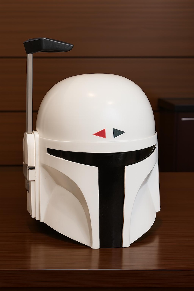 K&ouml;nnte beinhalten: Wei&szlig;er Mandalorianer-Helm mit schwarzen Akzenten, einschlie&szlig;lich eines T-f&ouml;rmigen Visiers und einer vertikalen Antenne. Der Helm hat rote und graue Dreiecksdetails und steht auf einer braunen Oberfl&auml;che.