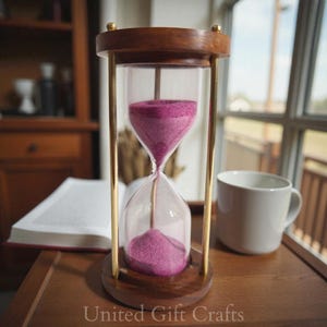 以下が含まれることがあります： 木製のフレームと真鍮のアクセントが付いた砂時計。ガラスのチャンバーにはピンク色の砂が入っています。白いマグカップと開いた本が木製のテーブルにあります。「United Gift Crafts」の文字が下にあります。