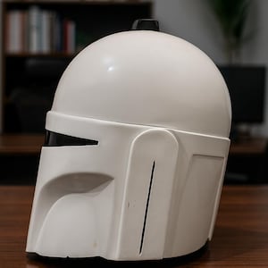 K&ouml;nnte beinhalten: Wei&szlig;er Mandalorianer-Helm mit schwarzem Visier und kleiner schwarzer Antenne. Der Helm besteht aus glattem, gl&auml;nzendem Material und hat ein markantes, eckiges Design. Er steht auf einer braunen Holzoberfl&auml;che.