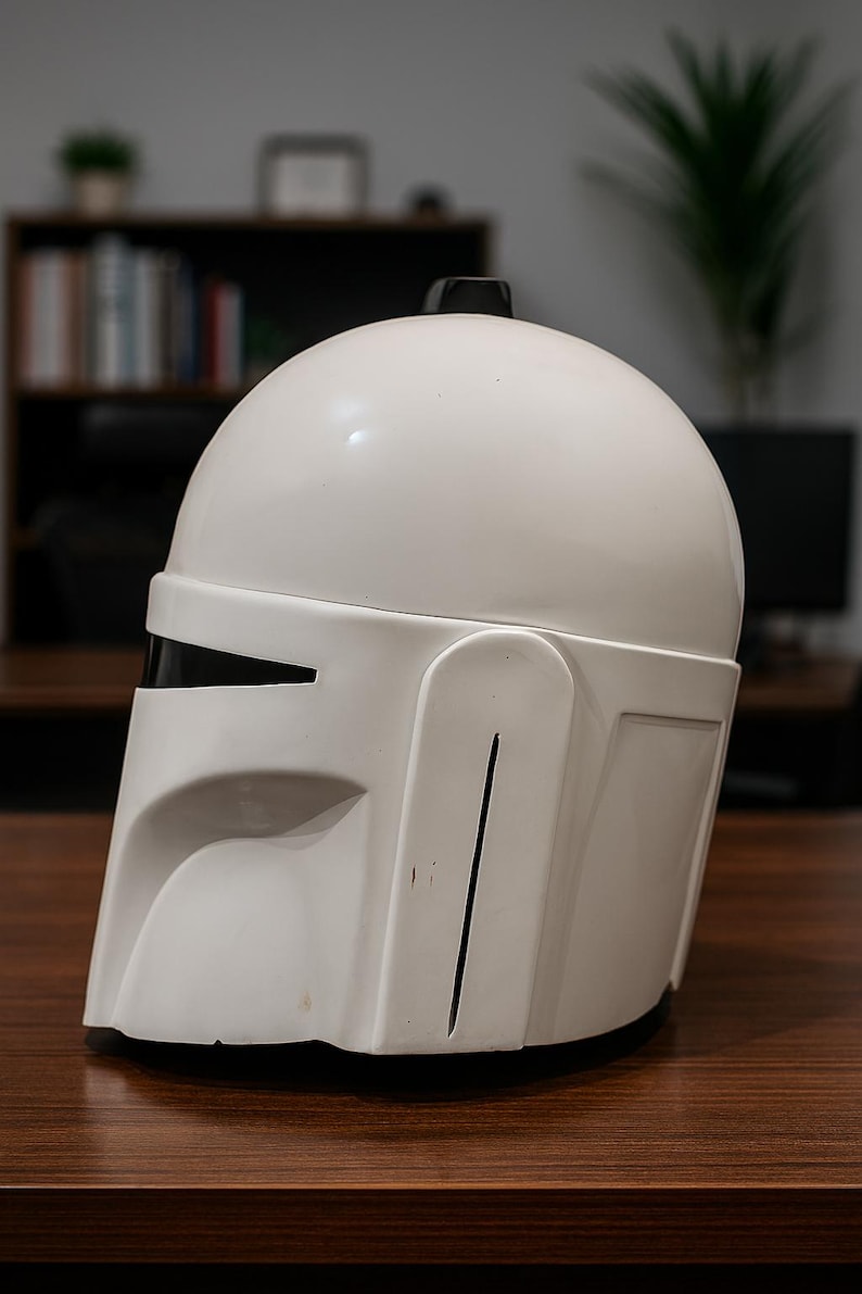 K&ouml;nnte beinhalten: Wei&szlig;er Mandalorianer-Helm mit schwarzem Visier und kleiner schwarzer Antenne. Der Helm besteht aus glattem, gl&auml;nzendem Material und hat ein markantes, eckiges Design. Er steht auf einer braunen Holzoberfl&auml;che.