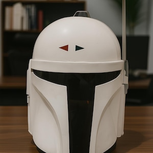 K&ouml;nnte beinhalten: Wei&szlig;er Mandalorianer-Helm mit schwarzem Visier und Akzenten. Der Helm hat einen schwarzen und grauen Entfernungsmesser und zwei kleine Dreiecke, eines rot und eines gr&uuml;n, auf der Stirn. Der Helm steht auf einer braunen Oberfl&auml;che.