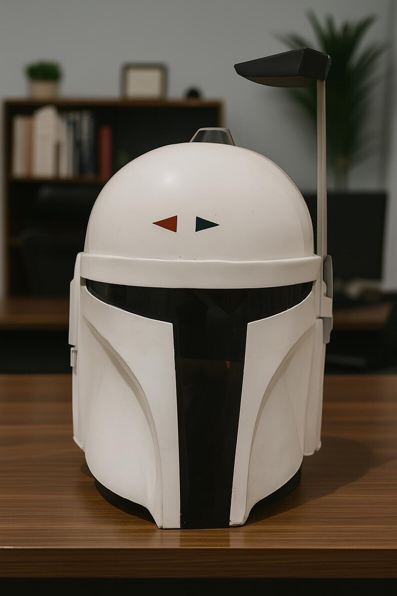 K&ouml;nnte beinhalten: Wei&szlig;er Mandalorianer-Helm mit schwarzem Visier und Akzenten. Der Helm hat einen schwarzen und grauen Entfernungsmesser und zwei kleine Dreiecke, eines rot und eines gr&uuml;n, auf der Stirn. Der Helm steht auf einer braunen Oberfl&auml;che.