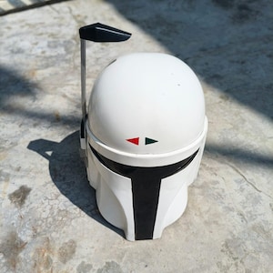 K&ouml;nnte beinhalten: Wei&szlig;er Mandalorianer-Helm mit schwarzem Visier und schwarzer Antenne. Der Helm hat rote und gr&uuml;ne Markierungen. Es ist eine Nachbildung des Helms aus der Star Wars-Serie. Der Helm liegt auf einer Betonoberfl&auml;che.
