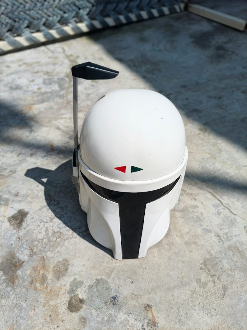 K&ouml;nnte beinhalten: Wei&szlig;er Mandalorianer-Helm mit schwarzem Visier und schwarzer Antenne. Der Helm hat rote und gr&uuml;ne Markierungen. Es ist eine Nachbildung des Helms aus der Star Wars-Serie. Der Helm liegt auf einer Betonoberfl&auml;che.