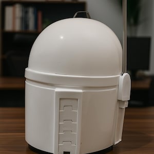 K&ouml;nnte beinhalten: Wei&szlig;er Mandalorianer-Helm mit abgerundeter Oberseite und rechteckigem Visier. Der Helm hat eine Reihe rechteckiger Details an der Seite und eine dunkelgraue Antenne. Der Helm steht auf einer braunen Holzoberfl&auml;che.