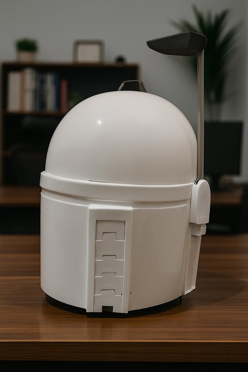 K&ouml;nnte beinhalten: Wei&szlig;er Mandalorianer-Helm mit abgerundeter Oberseite und rechteckigem Visier. Der Helm hat eine Reihe rechteckiger Details an der Seite und eine dunkelgraue Antenne. Der Helm steht auf einer braunen Holzoberfl&auml;che.