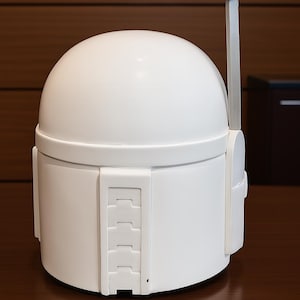 K&ouml;nnte beinhalten: Wei&szlig;er Mandalorianer-Helm mit abgerundeter Oberseite und rechteckigen Details an der Seite. Der Helm hat eine schwarze antennenartige Struktur, die sich von oben erstreckt. Der Helm wird auf einer dunkelbraunen Oberfl&auml;che pr&auml;sentiert.