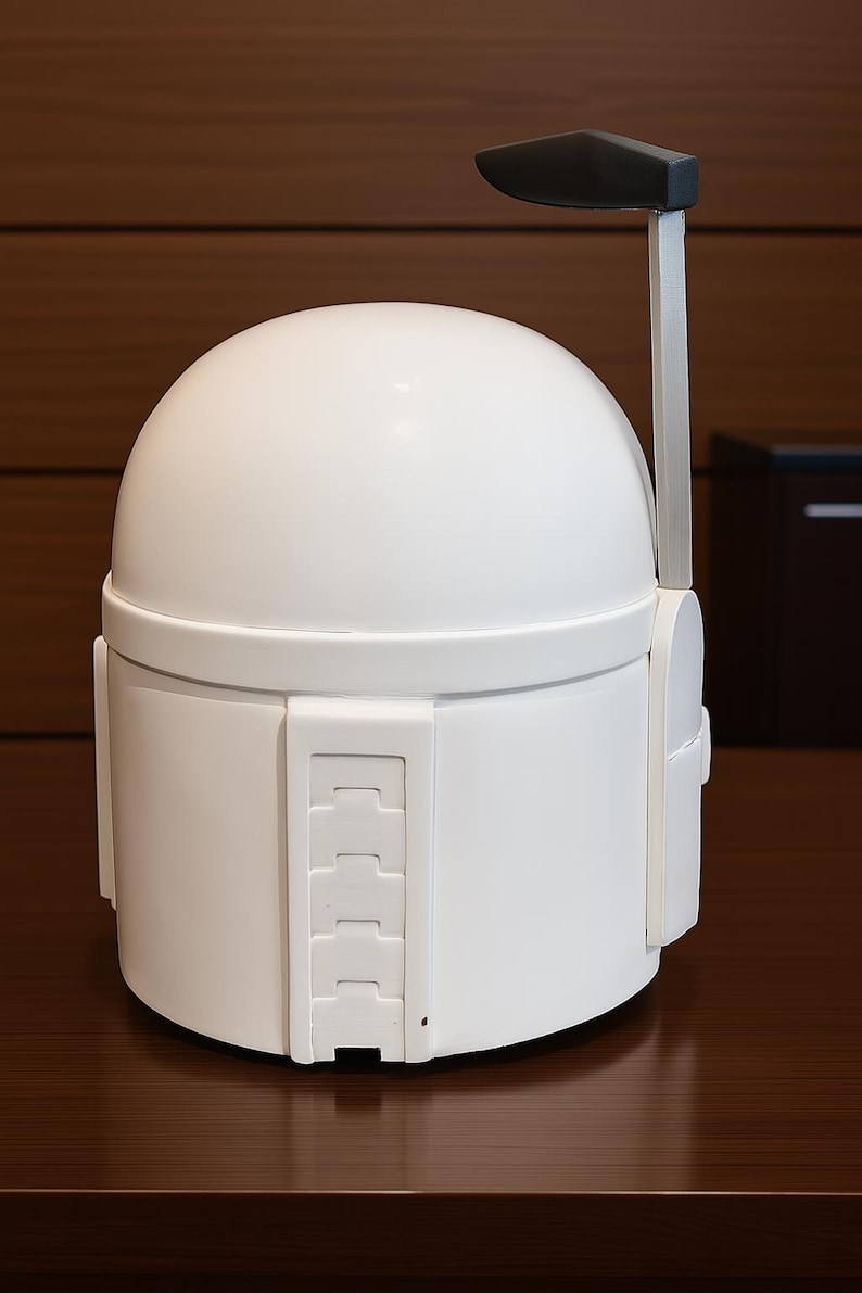 K&ouml;nnte beinhalten: Wei&szlig;er Mandalorianer-Helm mit abgerundeter Oberseite und rechteckigen Details an der Seite. Der Helm hat eine schwarze antennenartige Struktur, die sich von oben erstreckt. Der Helm wird auf einer dunkelbraunen Oberfl&auml;che pr&auml;sentiert.