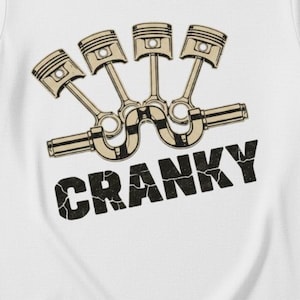 May include: White t-shirt featuring a graphic of four pistons and the word "CRANKY" in a distressed font. The pistons are beige and the text is black, creating a bold contrast on the white fabric. A casual, automotive-themed design.