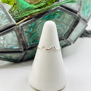 May include: A silver ring with a wavy design sits on a white display stand. The ring is made of thin wire and has a delicate, feminine look.