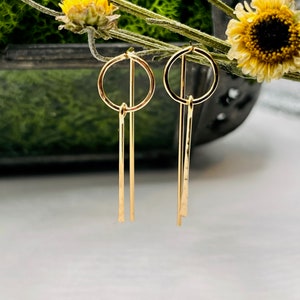 May include: A pair of gold earrings with a simple circle design and a long, thin dangle. The earrings are made of a smooth, shiny metal.