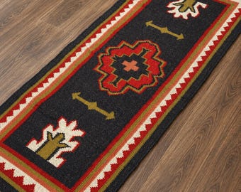 Puede incluir: Una alfombra rectangular tipo pasillero con fondo gris oscuro y un motivo geométrico central en rojo y naranja. El borde presenta un patrón en zigzag blanco y rojo con detalles en verde oliva. Flecos marrones adornan ambos extremos.