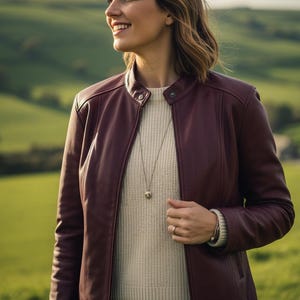 May include: A burgundy leather jacket with a round collar and a silver necklace. The jacket is worn over a cream-colored sweater. The background features green rolling hills.