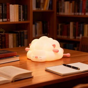 Puede incluir: Una lámpara suave y brillante con forma de nube y detalles en rosa, apoyada sobre una superficie de madera. La lámpara tiene una cara somnolienta y está rodeada de libros, creando un ambiente acogedor y relajante. Es un objeto decorativo y lindo.