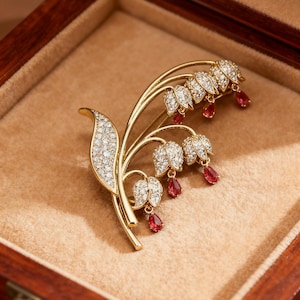 May include: A gold-toned brooch in the shape of a lily of the valley, set in a wooden box. The brooch features clear, sparkling stones and red teardrop-shaped accents. The brooch is a floral design.