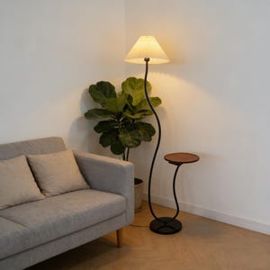 Könnte beinhalten: Eine Stehlampe mit plissiertem Lampenschirm und geschwungenem schwarzem Metallrahmen. Ein kleiner Beistelltisch mit runder Holzplatte und geschwungenem schwarzem Metallsockel. Ein graues Sofa und eine Topfpflanze befinden sich ebenfalls im Raum.