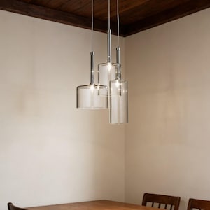 Op de afbeelding: Drie moderne hanglampen met heldere glazen kappen en chromen armaturen hangen aan het plafond. De kappen zijn cilindrisch en klokvormig en geven een warm licht. De lampen hangen boven een houten tafel.