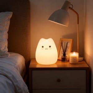 May include: A soft, glowing cat-shaped night light with a smiling face sits on a wooden nightstand. A white desk lamp and a lit candle are also on the nightstand. The scene is warm and inviting, with soft lighting.
