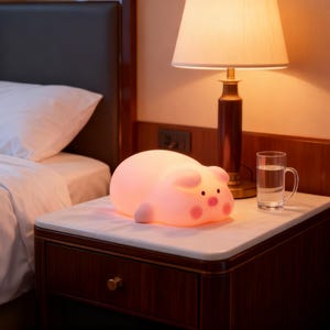 Puede incluir: Una suave luz nocturna con forma de cerdo rosa descansa sobre una mesita de noche con tapa de mármol. La lámpara emite un brillo cálido. Un vaso de agua y una lámpara con pantalla beige también están sobre la mesa. La mesa es de madera oscura.