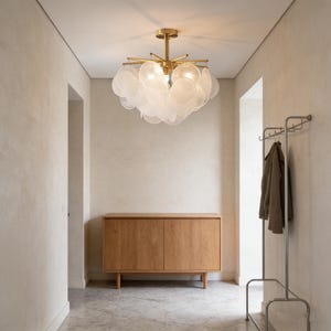 May include: A hallway featuring a contemporary chandelier, a wooden cabinet, and a metal coat rack. The chandelier has a gold finish with multiple lights and white, disc-shaped accents. The cabinet is made of light-coloured wood. A coat hangs on the rack.