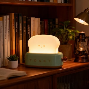 May include: A novelty night light shaped like a piece of toast in a toaster. The light is a soft yellow, with a smiling face on the toast. The toaster base is mint green with the word "TOAST" printed on it.
