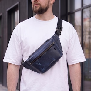 Pode incluir: Uma bolsa transversal de couro azul-marinho com acabamento liso e marmoreado azul claro, alça preta ajustável com fivela resistente, fecho de zíper superior e um pequeno remendo retangular de couro na parte inferior frontal.