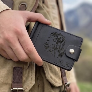 May include: Black leather wallet with a lion's head engraved on the front. The wallet has a snap closure and the initials "J.O. K." are visible. The wallet is held in a person's hand.