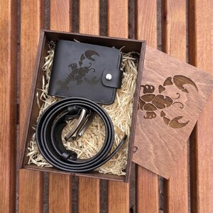 Pode incluir: Um conjunto de presente em couro preto com carteira e cinto com fivela prateada, apresentado numa caixa de madeira. A carteira e a tampa da caixa apresentam um design de escorpião. O conjunto inclui um cinto enrolado.