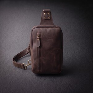 Pode incluir: Uma bolsa tiracolo de couro castanho escuro com um bolso frontal vertical com fecho, um compartimento principal com fecho e uma alça ajustável fixada com ferragens de metal em tom de latão, incluindo rebites e um mosquetão.