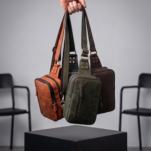 Pode incluir: Três bolsas tiracolo de couro nas cores castanho, verde-oliva e castanho escuro. Cada bolsa apresenta um formato retangular, um bolso frontal vertical com zíper, um compartimento principal com zíper e uma alça de ombro ajustável.