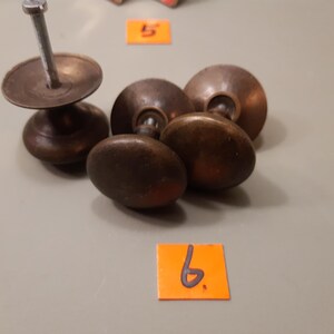 Może przedstawiać: Kolekcja antycznych gałek do szafek w odcieniach brązu i pojedyncza gałka ze śrubą. Gałki mają owalny kształt i ciemne, postarzane wykończenie. Przedmioty są ułożone na neutralnej powierzchni.
