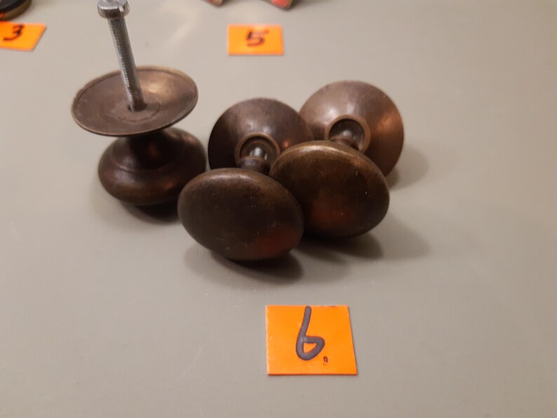 Może przedstawiać: Kolekcja antycznych gałek do szafek w odcieniach brązu i pojedyncza gałka ze śrubą. Gałki mają owalny kształt i ciemne, postarzane wykończenie. Przedmioty są ułożone na neutralnej powierzchni.
